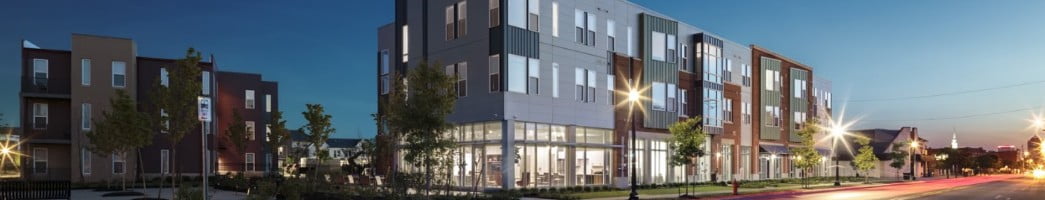 A three-story apartment building with a lower-level community space is lit up at dusk.
The building sits on a street corner and streetlights shine around the building, sprinkled amid several young trees.